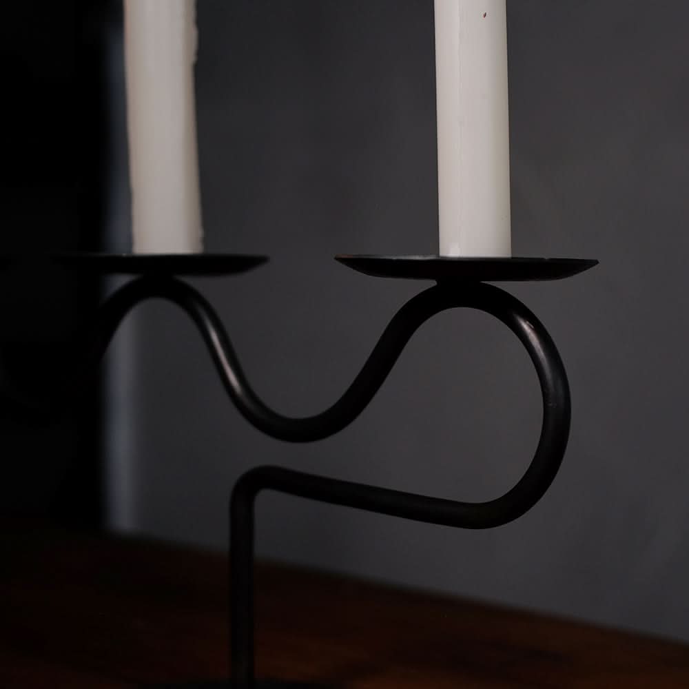 Black metal Three-Candle Holder with wavy design, holding two white candles on a wooden surface.